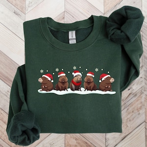 Op de afbeelding: Een donkergroen sweatshirt met een feestelijk ontwerp van vijf capybara's die kerstmanmutsen dragen en in een rij op een besneeuwde achtergrond staan.
