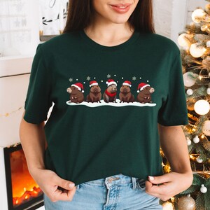 Op de afbeelding: Een donkergroen T-shirt met een feestelijk ontwerp van vijf capybara's die kerstmanmutsen dragen. De capybara's staan in een rij en staan op een besneeuwde ondergrond. Het ontwerp is gecentreerd op het shirt.