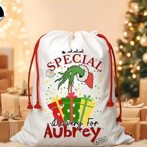 May include: White Christmas gift bag with red drawstring closure. The bag features a green Grinch hand design, the word "SPECIAL", and the text "Delivery For Aubrey". Presents and stars are also featured on the bag.