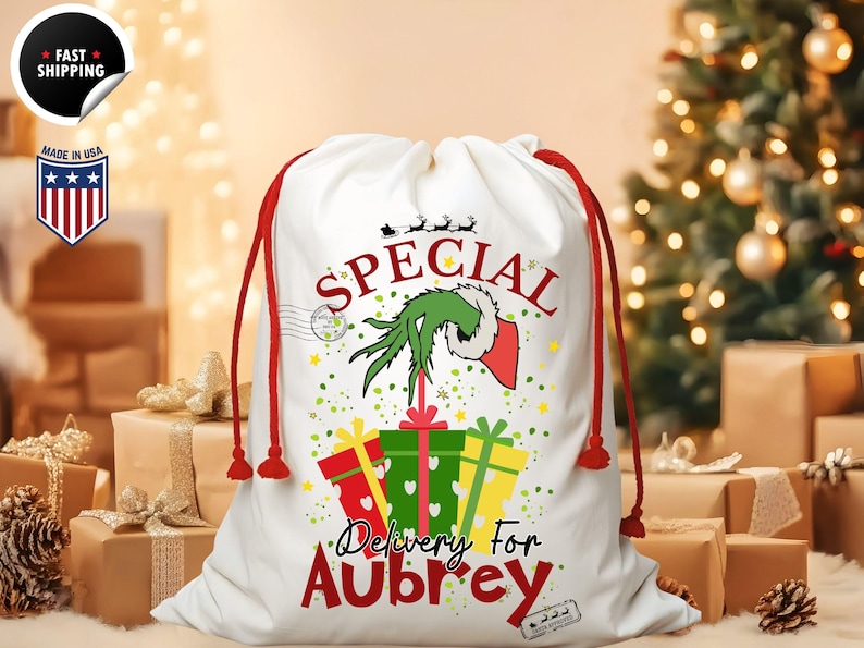 May include: White Christmas gift bag with red drawstring closure. The bag features a green Grinch hand design, the word "SPECIAL", and the text "Delivery For Aubrey". Presents and stars are also featured on the bag.