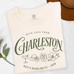 May include: A cream-colored t-shirt featuring dark green text that reads WITH LOVE FROM CHARLESTON and KATE'S BACHELORETTE - 2026. The graphic includes line art illustrations of a cocktail glass, a sun hat, flip-flops, and a magnolia flower.