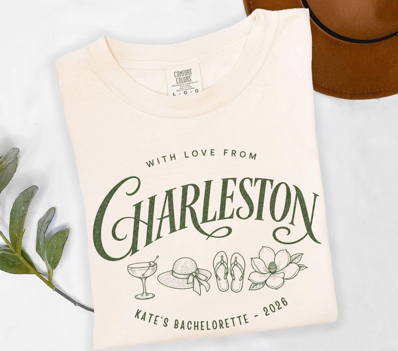 May include: A cream-colored t-shirt featuring dark green text that reads WITH LOVE FROM CHARLESTON and KATE'S BACHELORETTE - 2026. The graphic includes line art illustrations of a cocktail glass, a sun hat, flip-flops, and a magnolia flower.