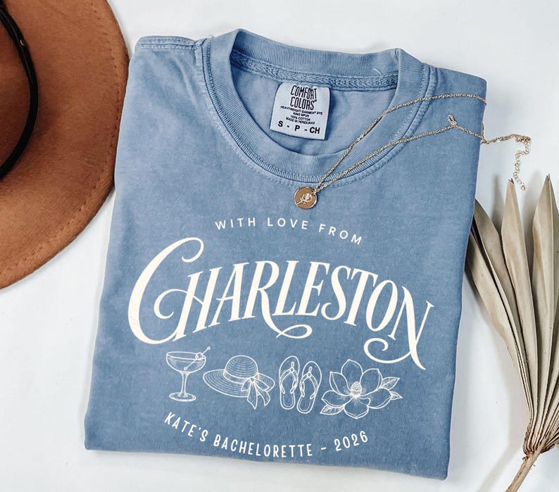 May include: A blue short-sleeved t-shirt featuring white text that reads WITH LOVE FROM CHARLESTON and KATE'S BACHELORETTE - 2026. The graphic includes line art of a cocktail glass, a sun hat, flip-flops, and a magnolia flower.