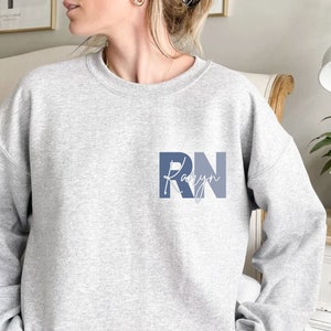 Puede incluir: Sudadera gris con un gr&aacute;fico azul y blanco que dice "RN Karyn".
