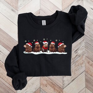 Op de afbeelding: Zwart sweatshirt met een kerstthema ontwerp met vijf capybara's die kerstmanmutsen dragen en in een rij staan op een besneeuwde achtergrond.