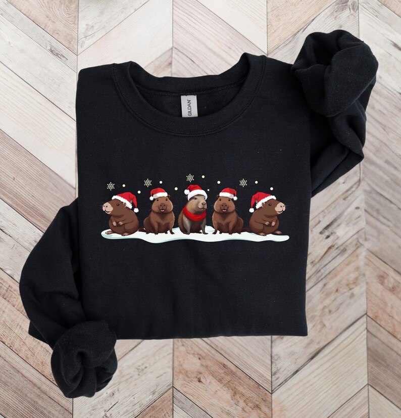 Op de afbeelding: Zwart sweatshirt met een kerstthema ontwerp met vijf capybara's die kerstmanmutsen dragen en in een rij staan op een besneeuwde achtergrond.