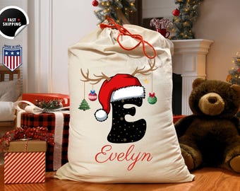 May include: A large, cream-colored Christmas gift sack with a red drawstring. The sack features a black letter "E" with a Santa hat and reindeer antlers. The name "Evelyn" is written in red script below the letter, with hanging ornaments.