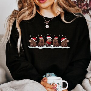 Op de afbeelding: Een zwart sweatshirt met een afbeelding van capibara's die kerstmanmutsen dragen. De capibara's zitten in een rij op een besneeuwde achtergrond. Het sweatshirt heeft een ronde hals en lange mouwen.