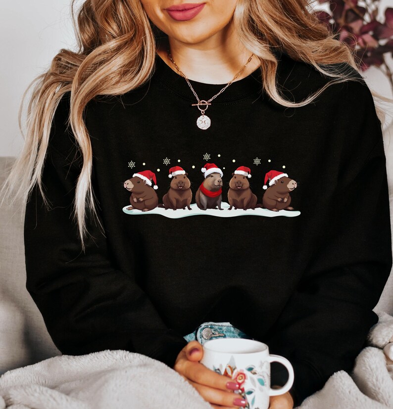 Op de afbeelding: Een zwart sweatshirt met een afbeelding van capibara's die kerstmanmutsen dragen. De capibara's zitten in een rij op een besneeuwde achtergrond. Het sweatshirt heeft een ronde hals en lange mouwen.