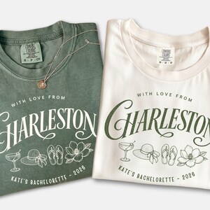 May include: A sage green t-shirt and a white t-shirt, both featuring the text 'WITH LOVE FROM CHARLESTON' and 'KATE'S BACHELORETTE - 2026' in a script font, accompanied by line art illustrations of a cocktail, a sun hat, flip-flops, and a magnolia.