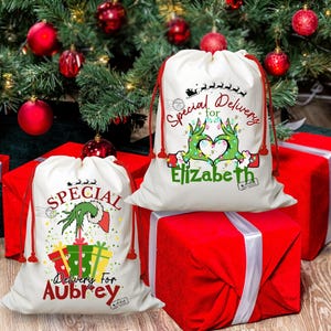 May include: Two white Christmas gift bags with red drawstrings, featuring Grinch-themed designs and the text "Special Delivery." One bag is personalized for "Aubrey," the other for "Elizabeth." The bags sit atop red wrapped gifts, with a Christmas tree in the background.