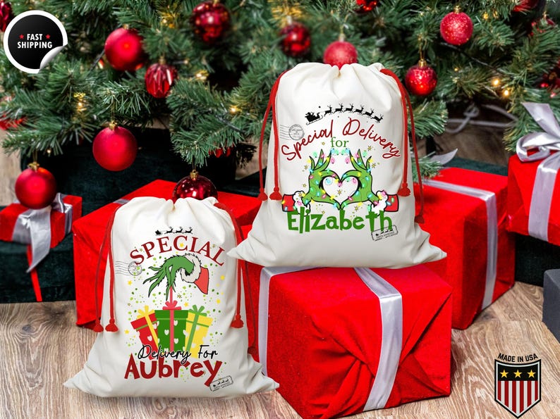 May include: Two white Christmas gift bags with red drawstrings, featuring Grinch-themed designs and the text "Special Delivery." One bag is personalized for "Aubrey," the other for "Elizabeth." The bags sit atop red wrapped gifts, with a Christmas tree in the background.