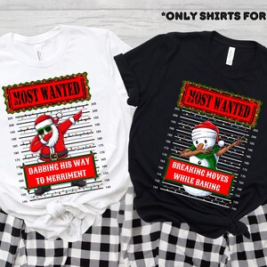 May include: Two t-shirts, one white and one black, with Christmas-themed graphics. The white shirt features Santa dabbing with the text "Most Wanted" and "Dabbing His Way to Merriment." The black shirt shows a snowman dabbing with the text "Breaking Moves While Baking."