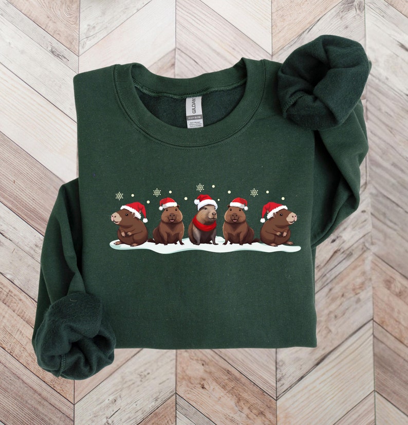 Op de afbeelding: Een donkergroen sweatshirt met een feestelijk ontwerp van vijf capybara's die kerstmanmutsen dragen en in een rij op een besneeuwde achtergrond staan.