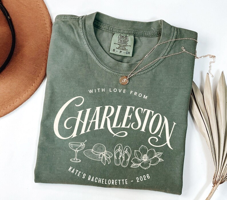 May include: An olive green crewneck t-shirt featuring white screen-printed text that reads "WITH LOVE FROM CHARLESTON" and "KATE'S BACHELORETTE - 2026". The graphic includes line art of a martini glass, a sun hat, flip-flops, and a magnolia flower.