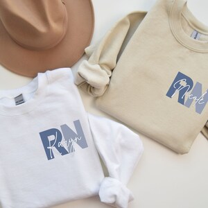 Puede incluir: Dos sudaderas blancas con texto azul y blanco. El texto de la sudadera de la izquierda dice "RN Kaylyn" y el texto de la sudadera de la derecha dice "RN Nicole".