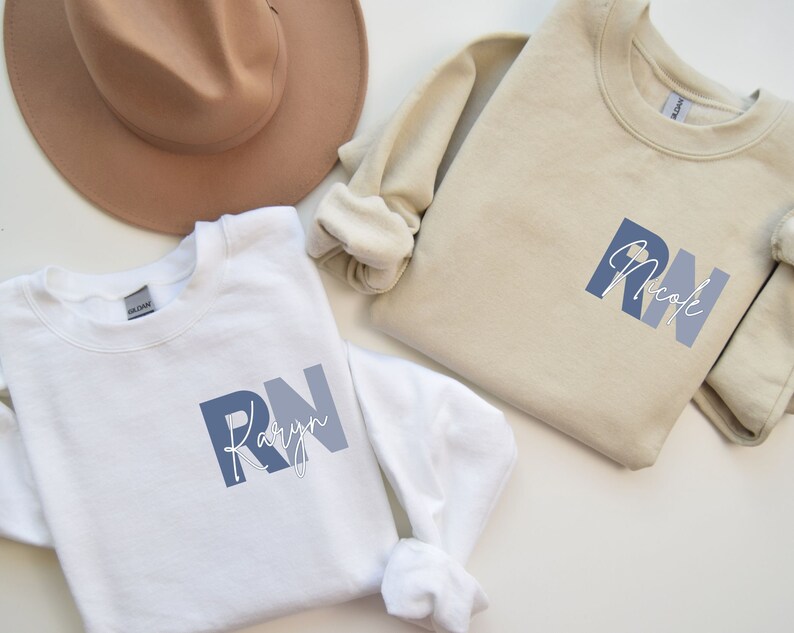 Puede incluir: Dos sudaderas blancas con texto azul y blanco. El texto de la sudadera de la izquierda dice "RN Kaylyn" y el texto de la sudadera de la derecha dice "RN Nicole".
