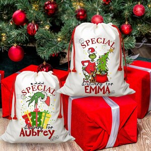 May include: Two white Christmas gift bags with red drawstrings, featuring a Grinch design and the words "Special Delivery for Aubrey" and "Special Delivery for Emma." The bags are placed on top of red wrapped gifts, with a Christmas tree and ornaments in the background.