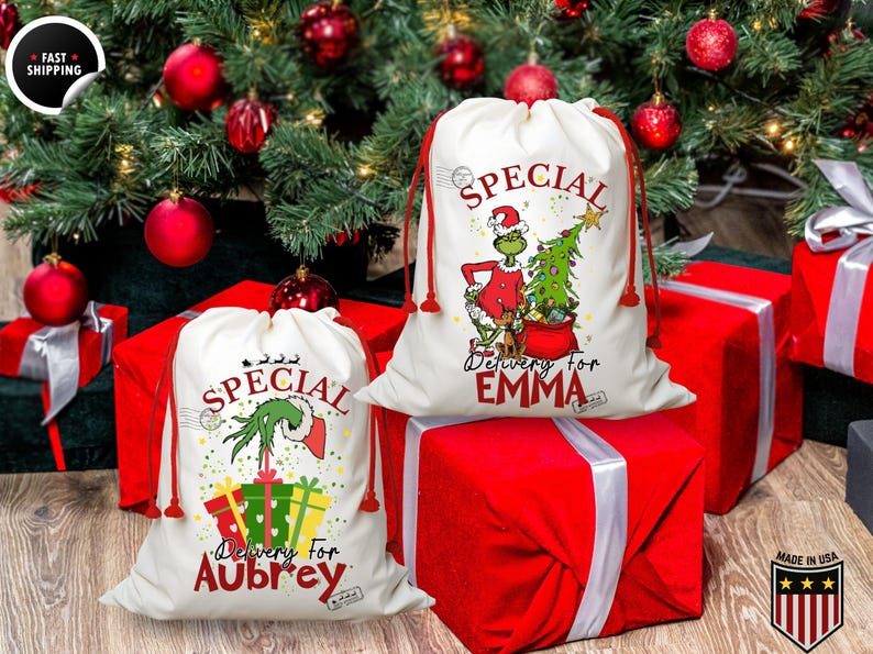 May include: Two white Christmas gift bags with red drawstrings, featuring a Grinch design and the words "Special Delivery for Aubrey" and "Special Delivery for Emma." The bags are placed on top of red wrapped gifts, with a Christmas tree and ornaments in the background.