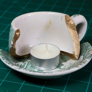 May include: A broken white teacup with gold accents is repurposed as a candle holder. A small white candle sits inside the cup on a green and white saucer.
