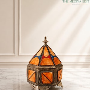 May include: A hexagonal decorative box with a brass frame and amber-colored resin panels. The lid features a pointed finial and alternating blue and red accents. The base is supported by three ornate brass feet. The text THE MEDINA EDIT MOROCCO is visible.