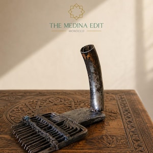 May include: A dark, weathered metal tool featuring a wide, multi-pronged comb-like head and a curved, tubular handle. The object displays signs of age and oxidation. The text THE MEDINA EDIT MOROCCO is visible above the object in the image.