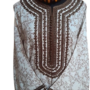May include: A white tunic with brown floral patterns and intricate brown embroidery around the neckline and sleeves. The tunic has a button-up front and is paired with a beaded necklace. The Medina Edit logo is in the top left corner.