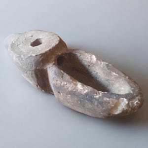 Könnte beinhalten: Eine kleine, graue Stein-Öllampe mit einem Loch oben für einen Docht. Die Lampe ist bootförmig und hat ein abgerundetes, ovales Becken.