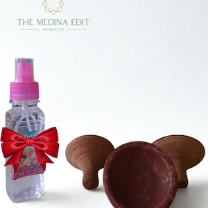 Puede incluir: Un frasco de spray transparente con tapa rosa y lazo rojo, junto a tres quemadores de incienso de arcilla marrón. El texto "THE MEDINA EDIT MOROCCO" está en la parte superior.