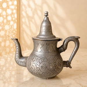May include: A silver-toned metal teapot featuring an intricate hand-engraved pattern of geometric shapes and circular motifs. The teapot has a curved spout, a sturdy handle, and a pointed lid with a small knob. Text reads THE MEDINA EDIT MOROCCO.