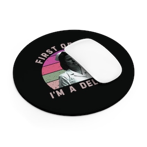 May include: A black round mouse pad with a retro sunset design and the text "First of all I'm a deli..." in pink. A white computer mouse is on top of the mouse pad.