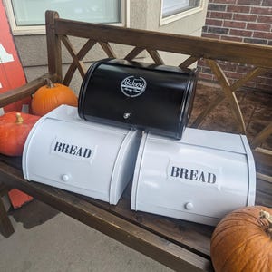 May include: A black metal bread box with a Bakery logo sits atop two white bread boxes, each labeled "BREAD" in black. The boxes are on a wooden bench with pumpkins, and a red and white sign in the background.