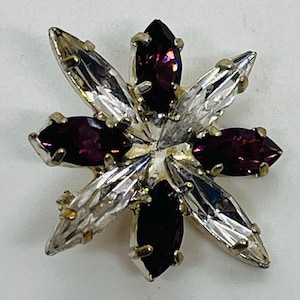 May include: A vintage flower-shaped brooch with alternating clear and deep purple crystal-like stones. The brooch has a silver-tone metal setting and is designed to resemble a star or flower. The crystals are faceted, catching the light.