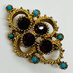 May include: A vintage gold brooch with a floral design. The brooch features four large, round, deep red gemstones and several smaller turquoise-coloured stones. The brooch has an ornate, textured gold metalwork design.