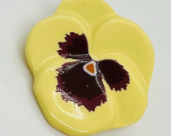 May include: A yellow ceramic pansy flower with a dark burgundy center and a small orange and white detail. The flower has a glossy finish and is set against a white background. This decorative piece is likely a brooch or a decorative element.