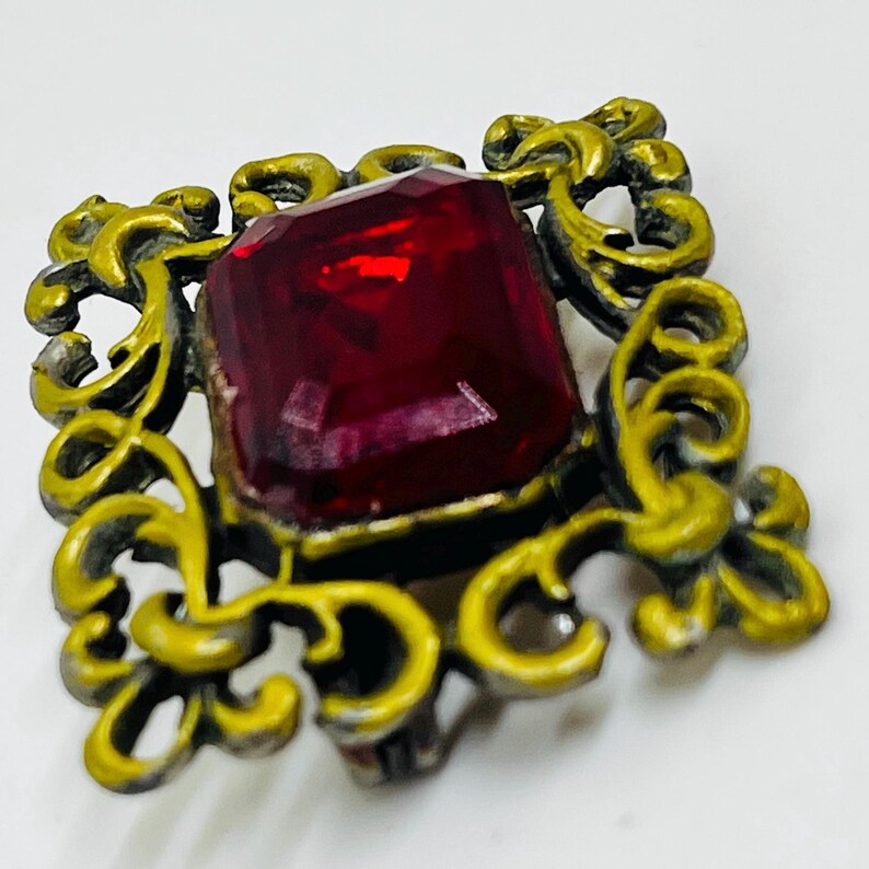 May include: A square brooch featuring a large, faceted, deep red gemstone set in the centre. The stone is surrounded by an ornate, openwork metal frame with a distressed, matte yellow finish over a dark base, creating a textured appearance.