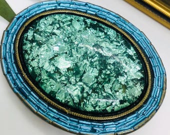 May include: A vintage oval-shaped brooch with a green, iridescent, mosaic-like center surrounded by a gold-colored frame and a blue enamel border.