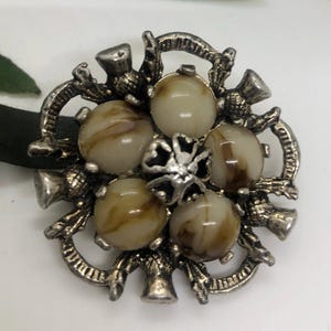 May include: A silver brooch with a floral design featuring six brown and white swirl patterned stones. The brooch is surrounded by a decorative border of thistle motifs.