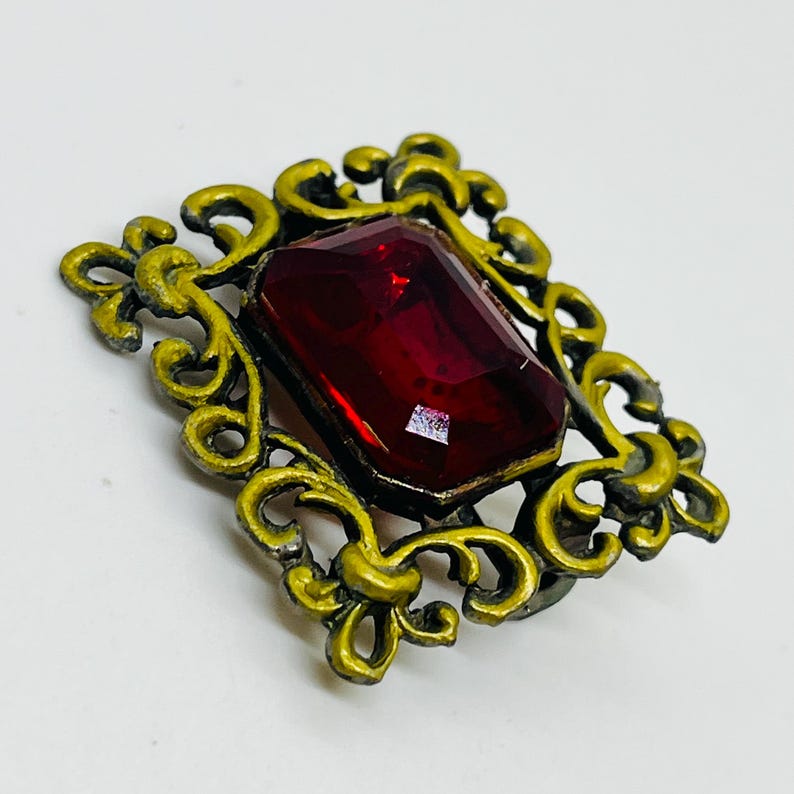 May include: A rectangular brooch featuring a large, faceted, deep red gemstone set within an ornate, antiqued gold-tone metal frame. The metalwork displays intricate, swirling scroll patterns that surround the central stone on all four sides.