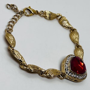 May include: Gold-toned bracelet featuring a large, heart-shaped red gemstone with a halo of small, clear crystals. The bracelet has a textured, leaf-like design and an adjustable chain.