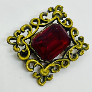 May include: A rectangular brooch featuring a large, faceted, deep red gemstone set within an ornate, scrollwork frame finished in a distressed, metallic gold paint with visible dark grey undertones along the edges and recessed areas.