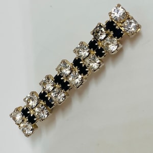 May include: A silver and black rhinestone brooch with a linear design. The brooch is made up of alternating black and clear rhinestones.