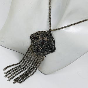 May include: A silver-toned necklace with a large, ornate pendant featuring a floral design and small red gemstones. The pendant has dangling chains. The necklace is displayed on a white mannequin bust.