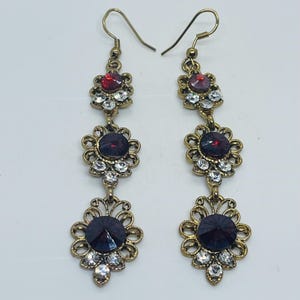 May include: A pair of long drop earrings featuring three tiered, filigree-style brass settings. Each tier contains a central round dark red crystal surrounded by smaller clear white rhinestones. The earrings hang from simple brass ear wires.
