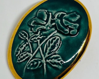 May include: An oval-shaped ceramic brooch with a gold rim. The brooch features a raised, white design of a single rose with leaves on a teal green background.