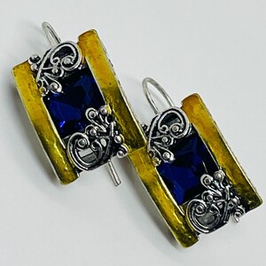 May include: A pair of rectangular earrings featuring a central deep blue faceted stone flanked by textured yellow metal borders. The design includes intricate silver-toned filigree scrollwork accents and a curved wire hook for attachment.