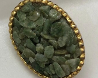 May include: Oval-shaped vintage brooch with a gold-tone beaded border. The center of the brooch is filled with small, irregularly shaped green stones, creating a textured surface. The brooch has a classic, elegant design.