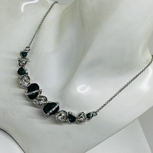 May include: A silver-toned necklace with a series of alternating dark green and clear crystal-like stones. The stones are set in a curved design, creating a unique and elegant look. The necklace is displayed on a white mannequin.