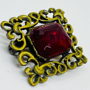 May include: A square brooch featuring a large, faceted, deep red rectangular gemstone set within an ornate, scrollwork frame finished in a distressed, matte yellow-gold tone. The metalwork displays intricate, swirling patterns surrounding the central stone.