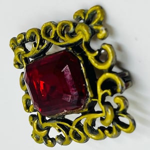May include: A square brooch featuring a central faceted deep red gemstone set within a decorative frame of yellow-painted metal with intricate scrollwork patterns. The metal frame displays a dark, weathered finish beneath the yellow paint.